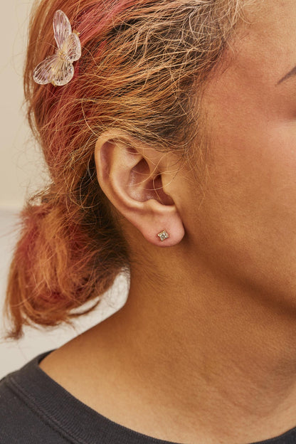 Close-up side view of a model wearing yellow metal stud earrings with square-shaped white gemstones in a four-prong setting.
