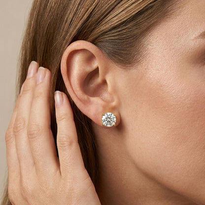 Close-up side view of a model wearing round white gemstone stud earrings with yellow metal 4-prong settings.