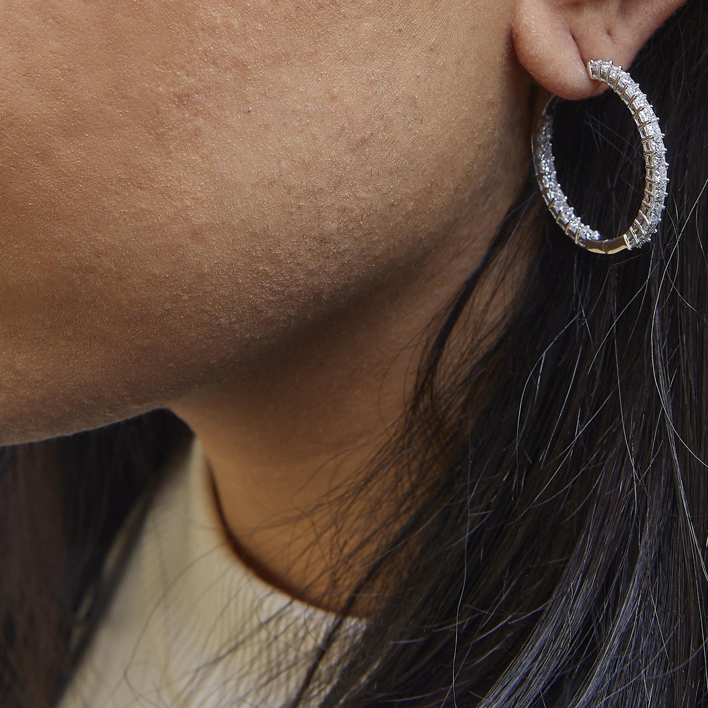 Close-up side view of a white metal hoop earring encrusted with multiple princess-shaped white gemstones in a shared prong setting, worn on a model.