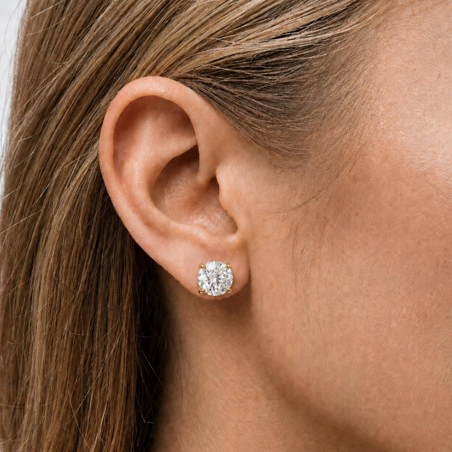 Close-up side view of a model wearing a yellow metal round stud earring with one white gemstone in a 4-prong setting.