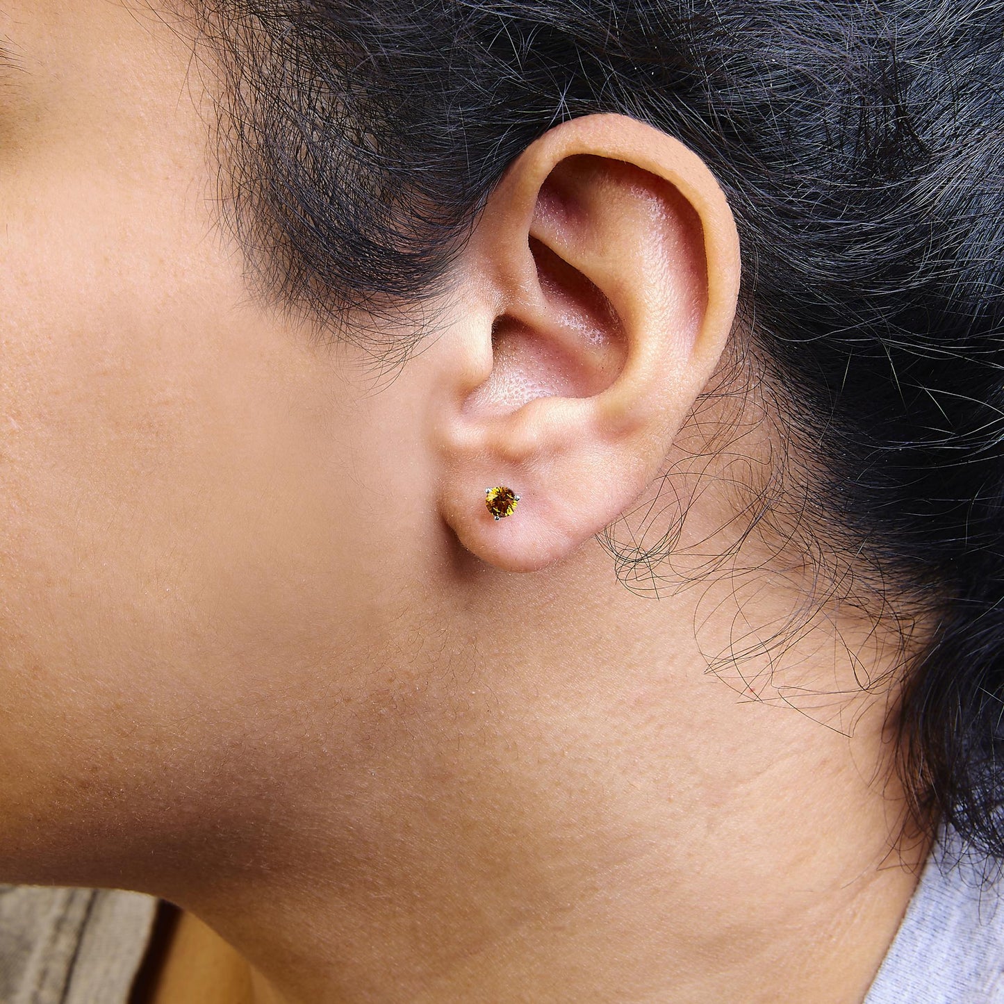Close-up side view of white metal stud earring with round orange gemstone in 3-prong setting, worn on a model's ear.