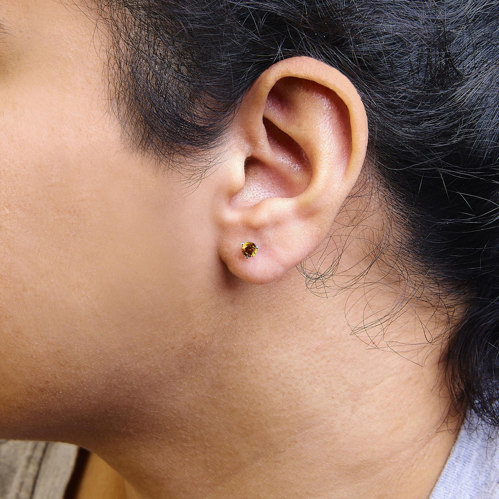 Close-up side view of white metal stud earring with round orange gemstone in 3-prong setting, worn on a model's ear.