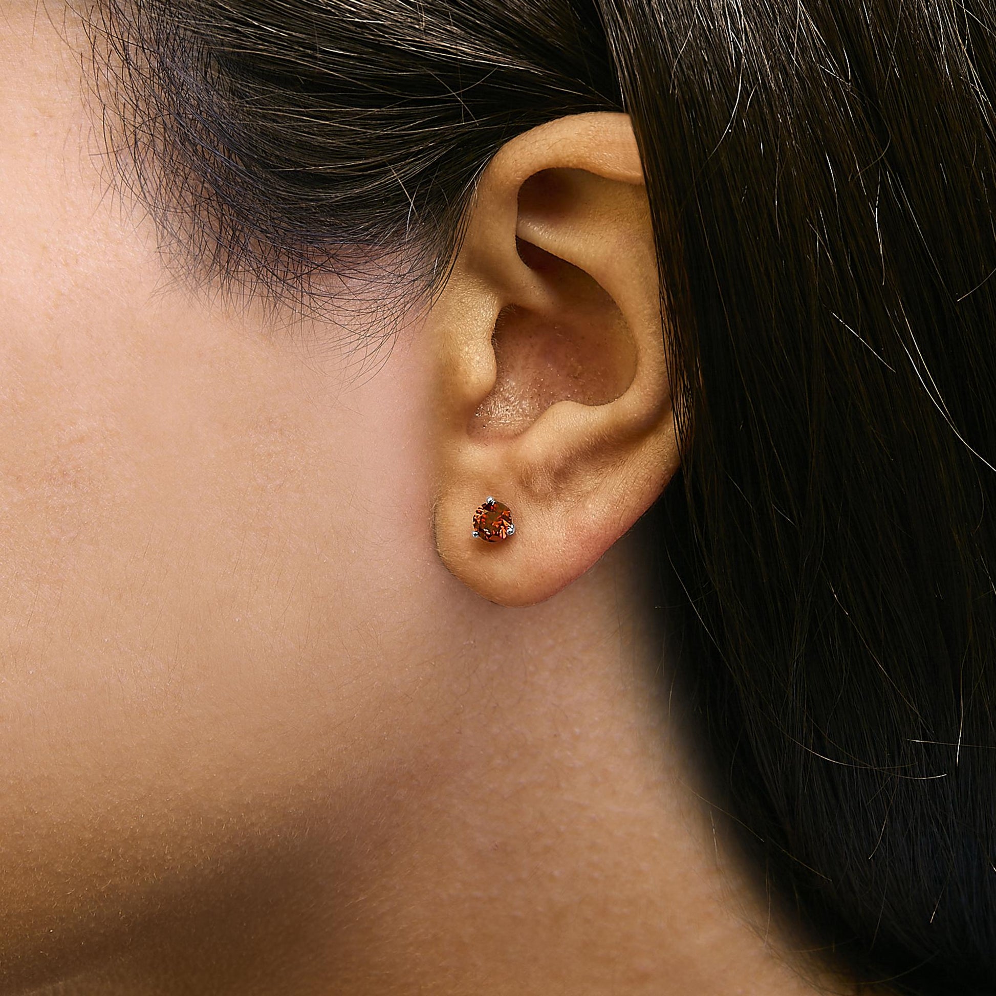 Close-up side view of a white metal stud earring with a round red gemstone in a three-prong setting, shown worn on a model's ear.