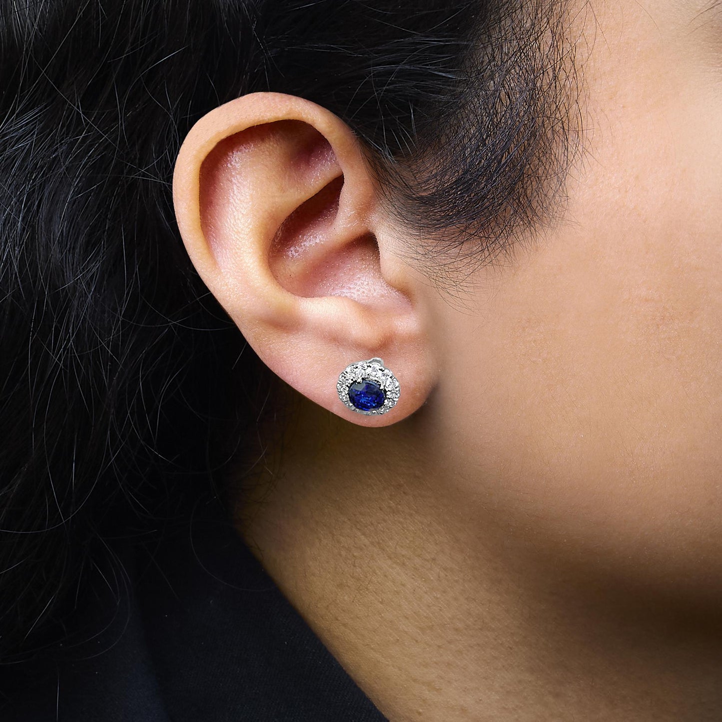 Close-up side view of a white metal stud earring worn on a model, featuring a central round blue gemstone surrounded by smaller round white gemstones in a halo design.