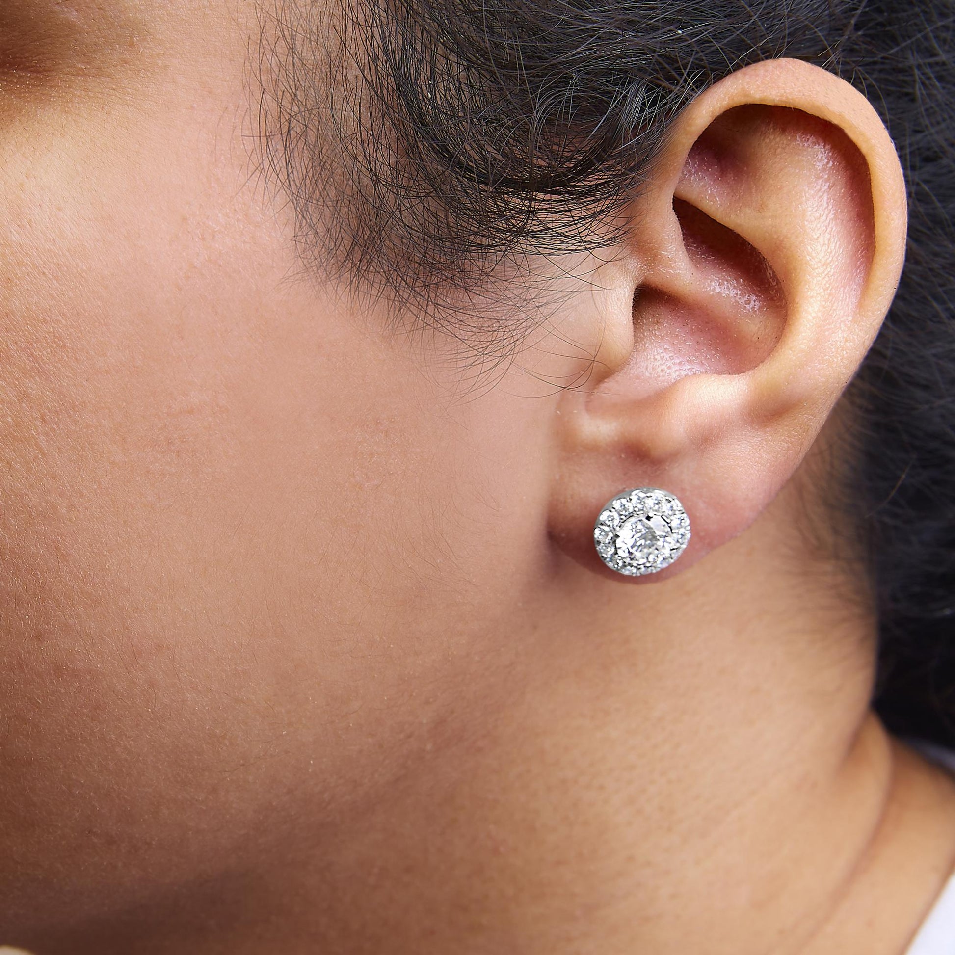 Close-up side view of white metal stud earrings with round white gemstones in prong settings, worn on a model.