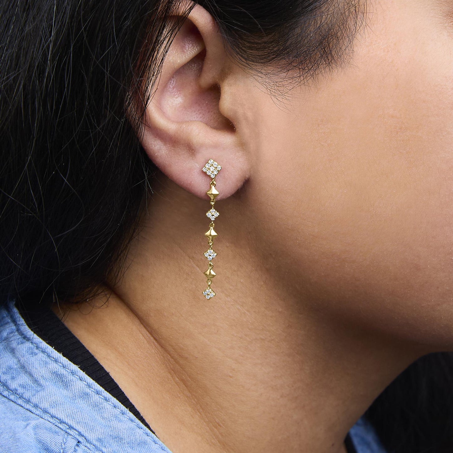 Close-up side view of a yellow metal drop earring on a model's ear, featuring multiple round white gemstones in prong settings.