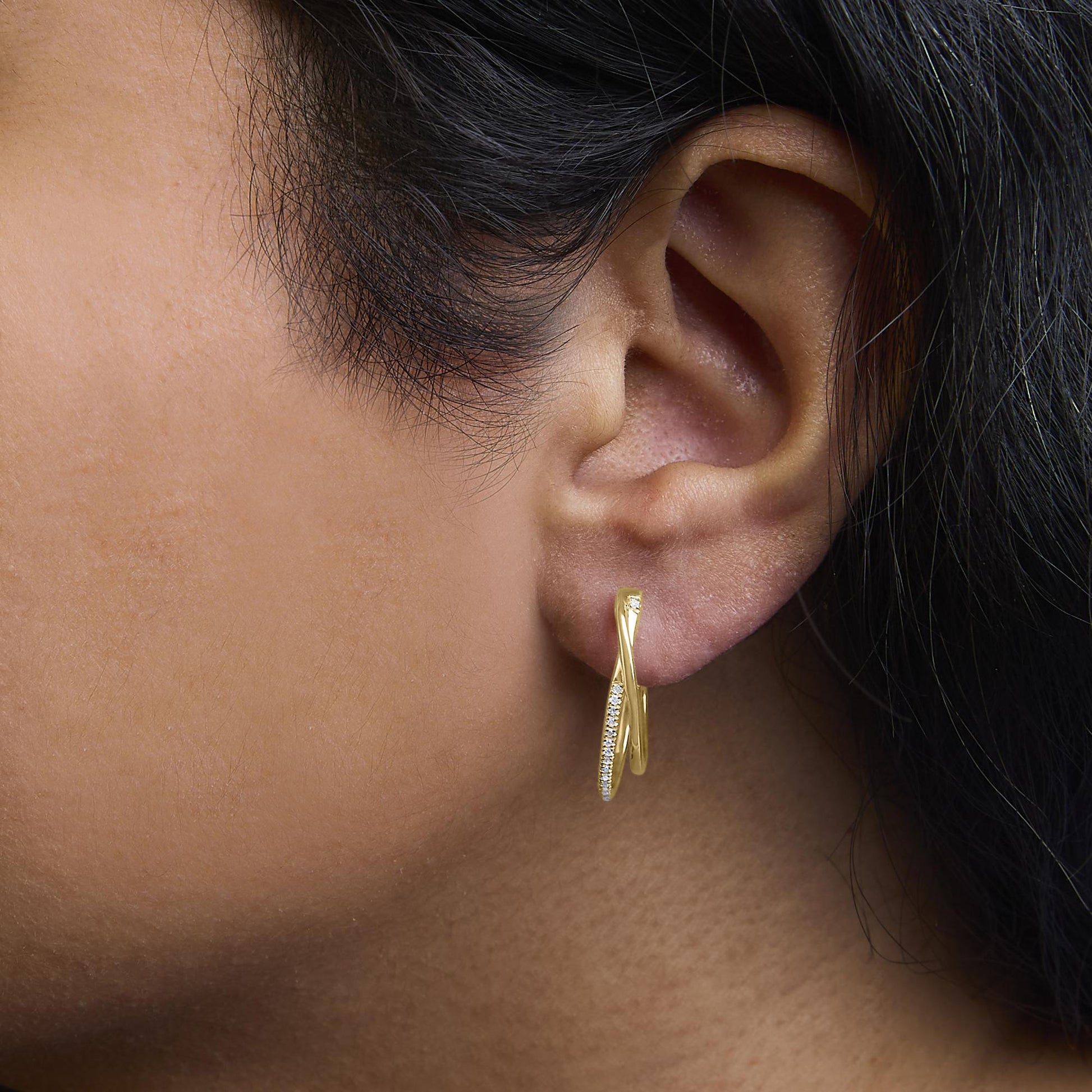 Close-up side view of a yellow metal earring worn on a model's ear, featuring two crossed loops with one loop adorned with small round white gemstones in a prong setting.