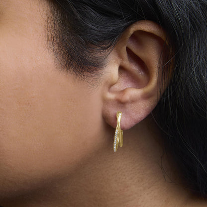 Close-up side view of a yellow metal earring worn on a model's ear, featuring two crossed loops with one loop adorned with small round white gemstones in a prong setting.