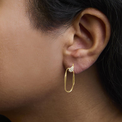 Close-up side view of yellow metal earrings with a twisted oval hoop and a cluster of round white gemstones in a pave setting, worn on a model.
