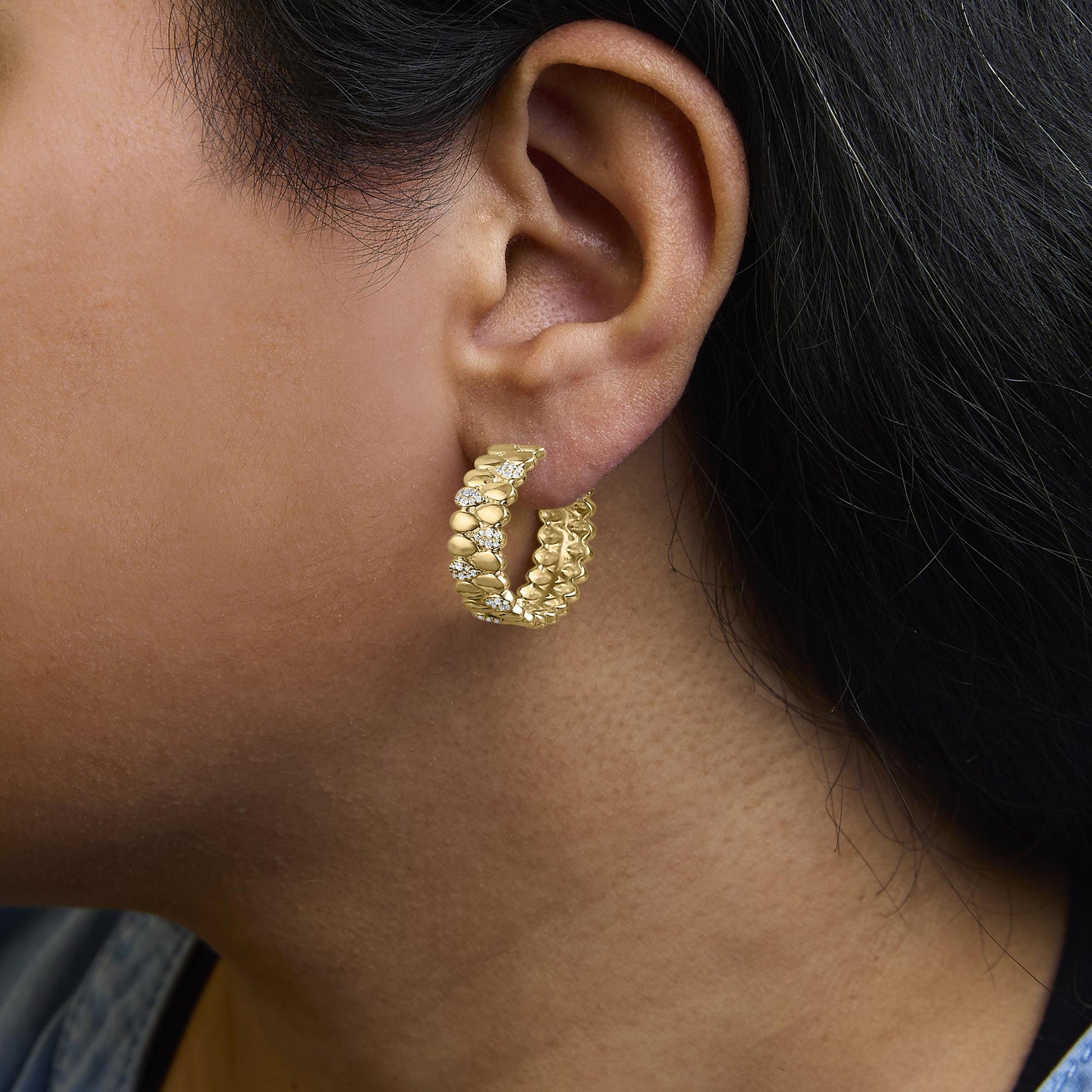 Close-up side view of a yellow metal hoop earring on a model's ear, featuring round white gemstones in prong and pave settings.