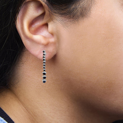 Close-up side view of a white metal earring on a model's ear, featuring a linear arrangement of round black gemstones in prong settings.