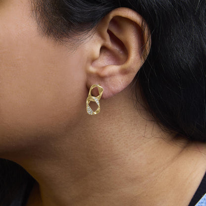 Close-up side view of a model wearing yellow metal earrings featuring intertwined loops adorned with white round gemstones in prong settings.