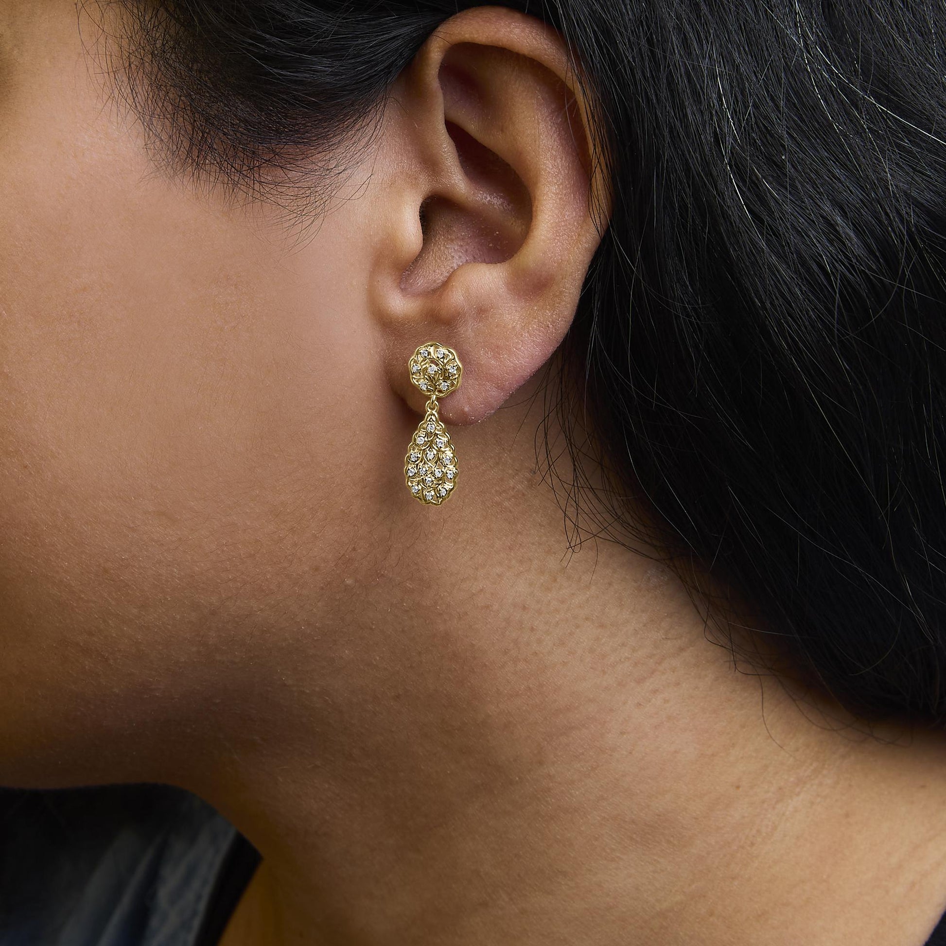 Close-up side view of a model wearing yellow metal earrings with round white gemstones in prong settings, featuring a floral-shaped stud and a teardrop-shaped dangling element.