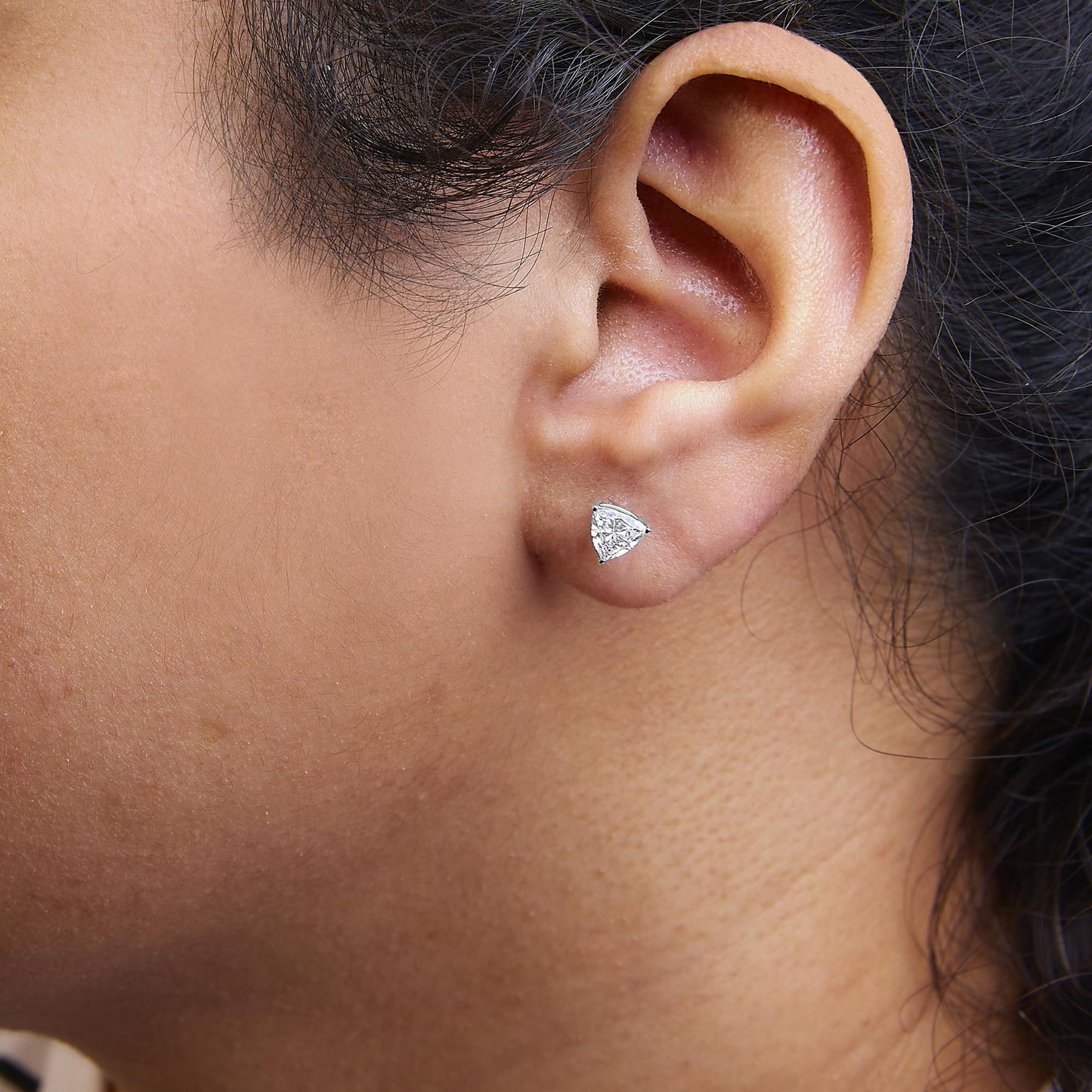 Close-up angled side view of a white metal earring with a trillion-shaped white gemstone in a 3-prong setting, worn on a model's ear.