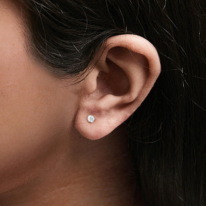Close-up side view of a white metal stud earring with a round white gemstone in a 3-prong setting worn on a model's ear.
