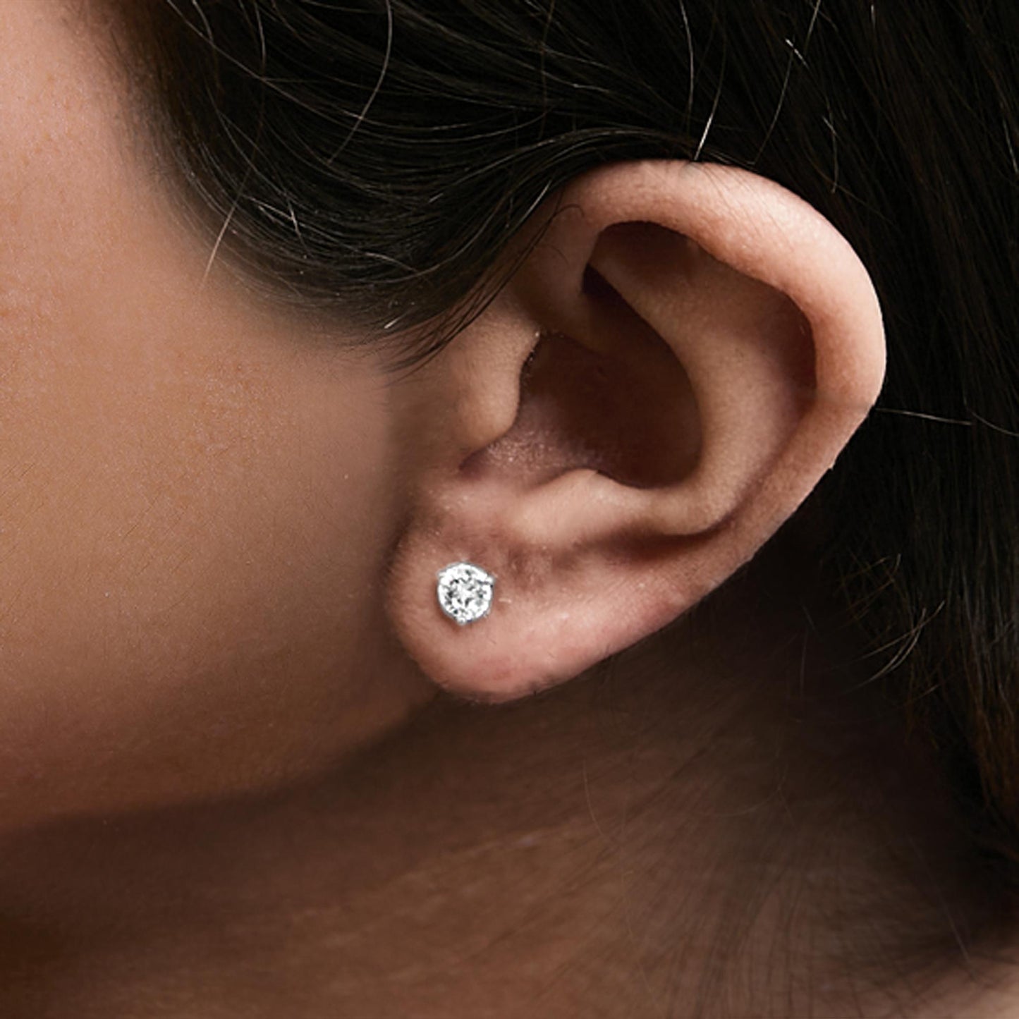 Close-up side view of a white metal stud earring with a round white gemstone in a three-prong setting, worn on a model's ear.