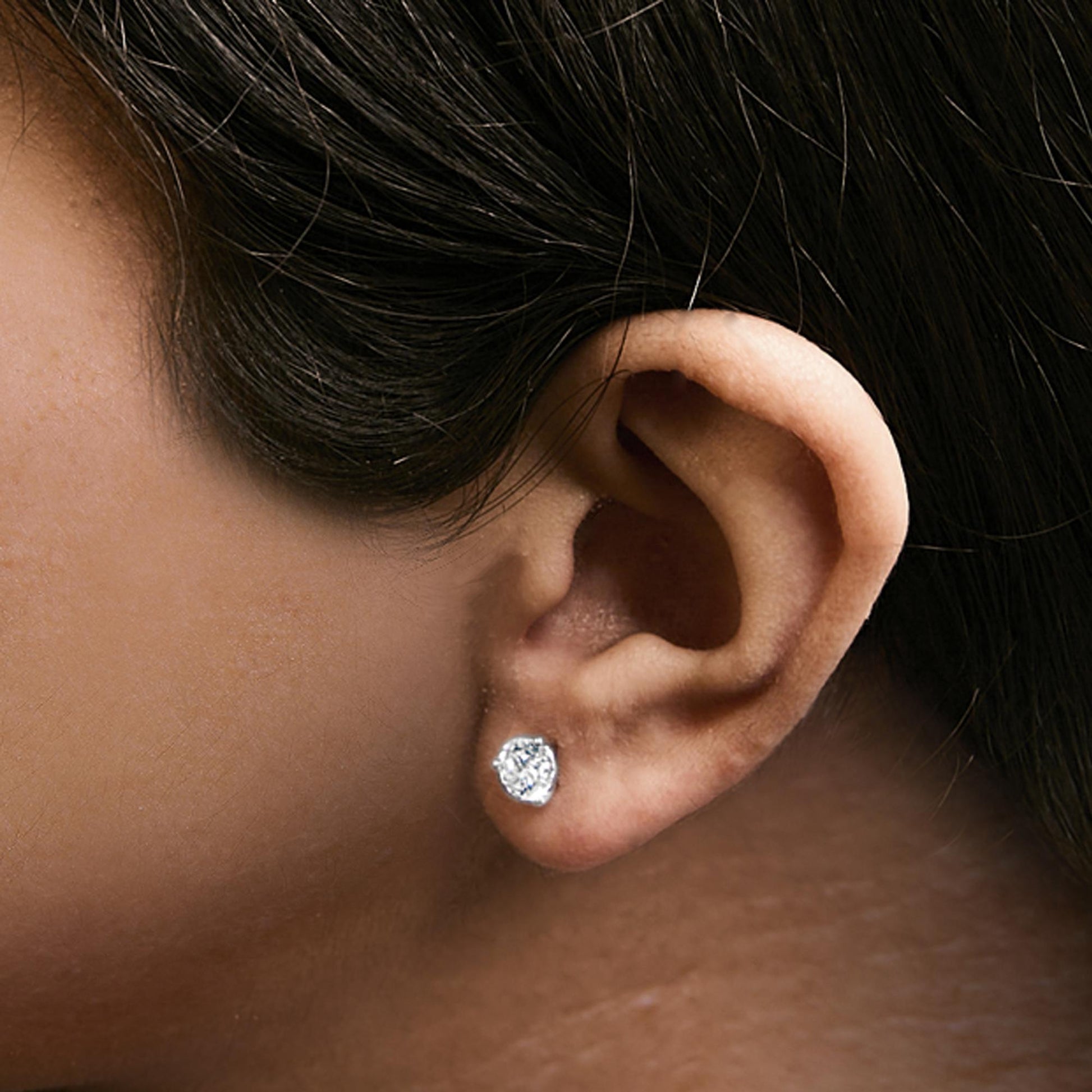 Close-up side view of an ear on a model wearing a white metal stud earring with a round white gemstone in a three-prong setting.