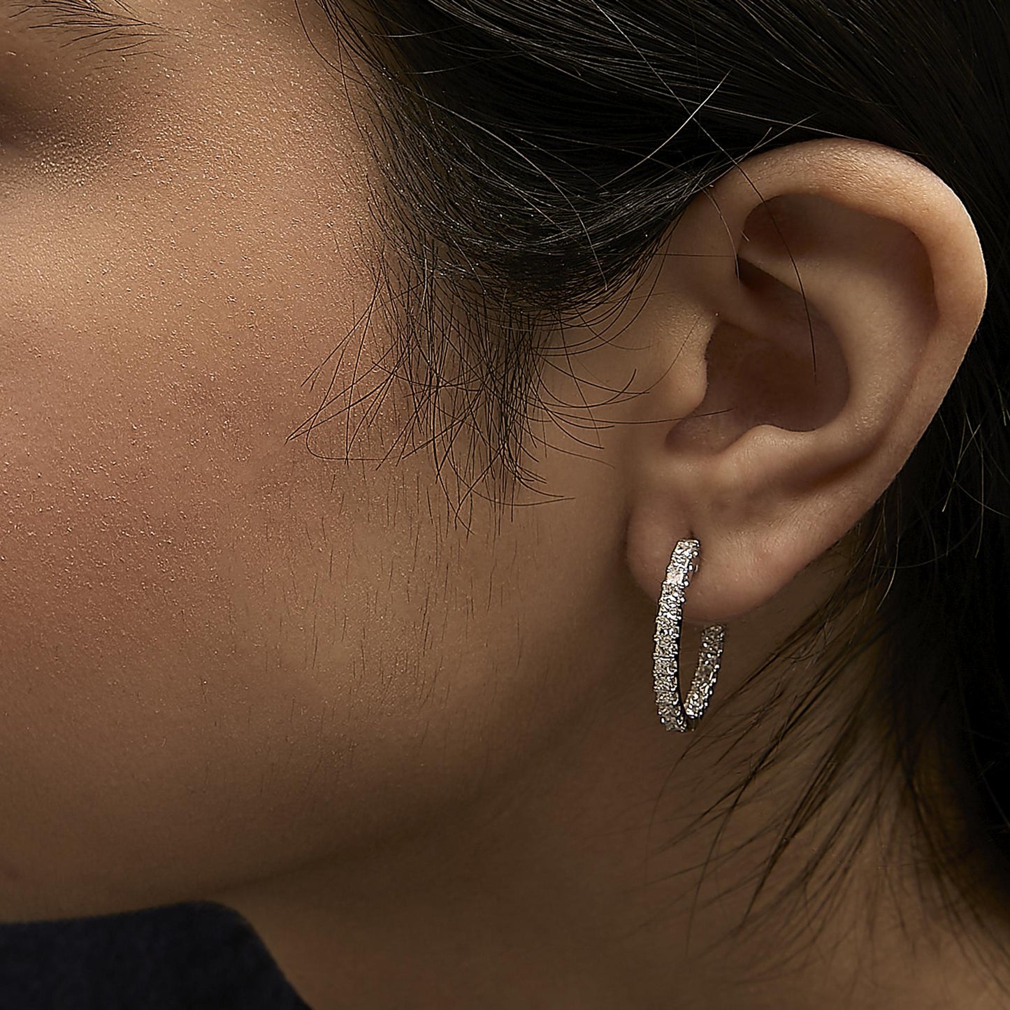 Close-up side view of a model wearing a white metal hoop earring encrusted with multiple princess-shaped white gemstones in a prong setting.