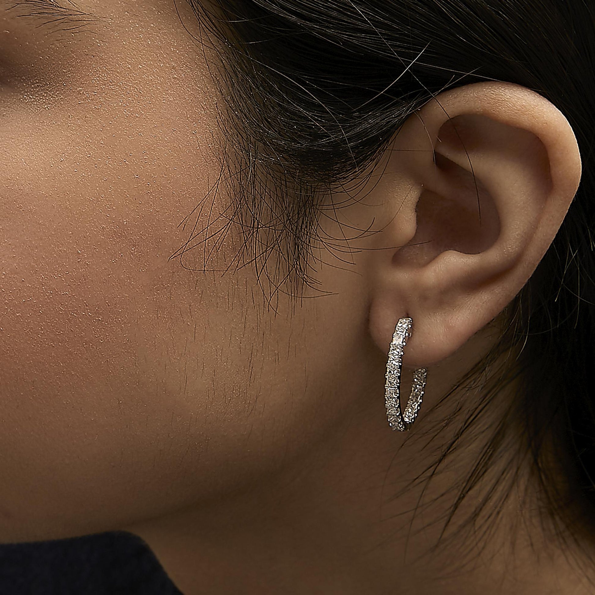 Close-up side view of a model wearing a white metal hoop earring encrusted with multiple princess-shaped white gemstones in a prong setting.