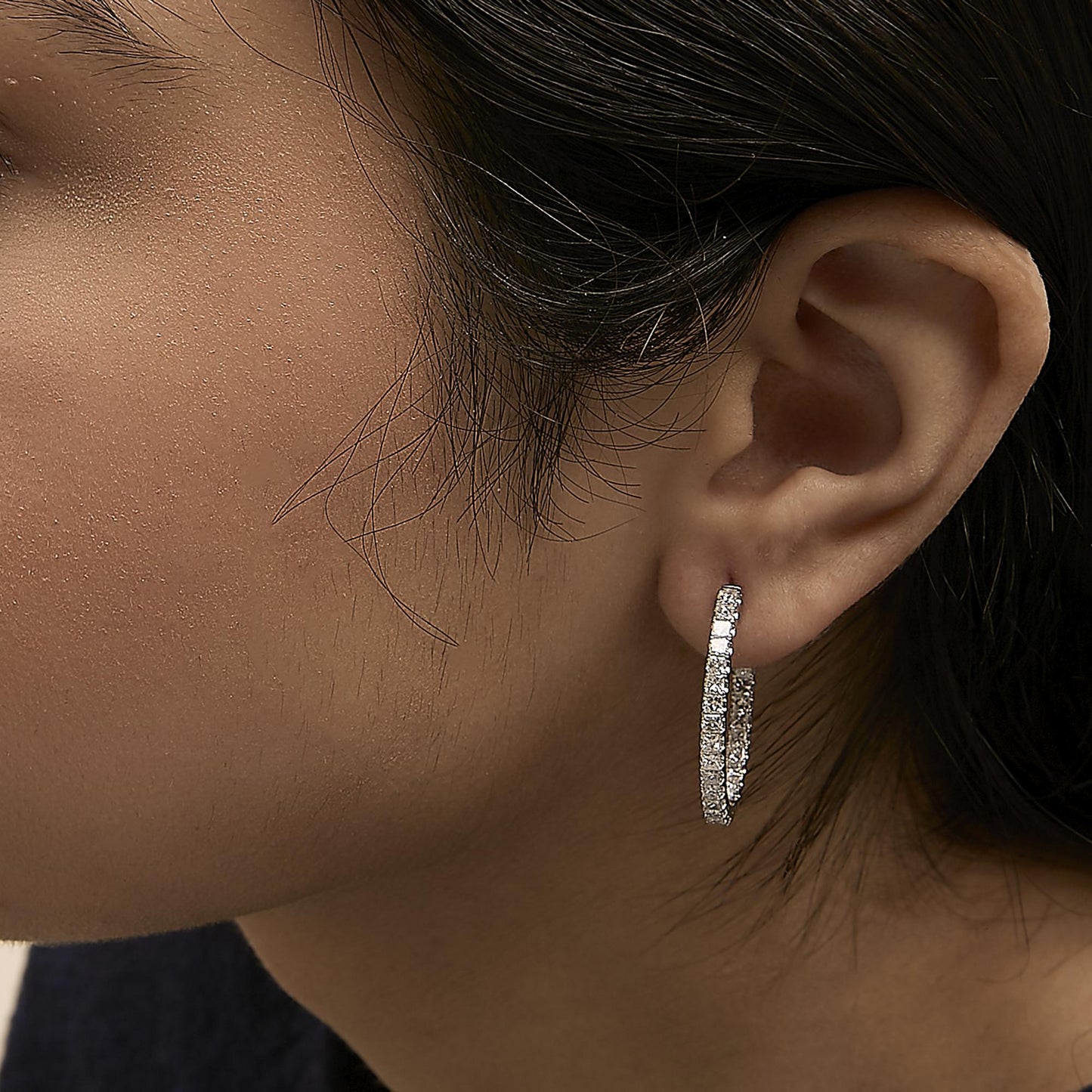 Close-up side view of a model wearing white metal hoop earrings with white princess-shaped gemstones in prong settings.