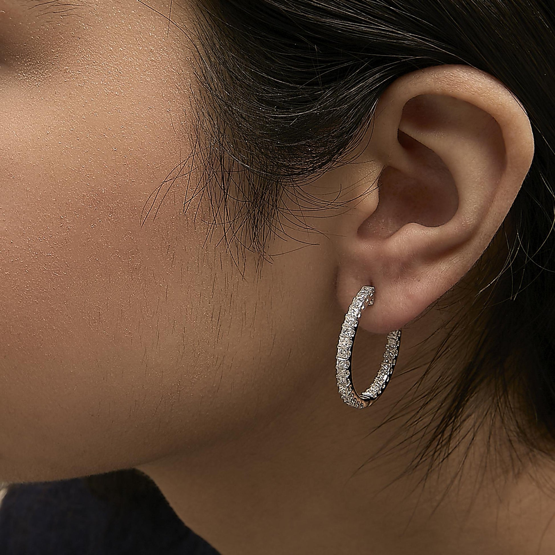 Close-up side view of a model wearing white metal hoop earrings encrusted with prong-set princess-shaped white gemstones.