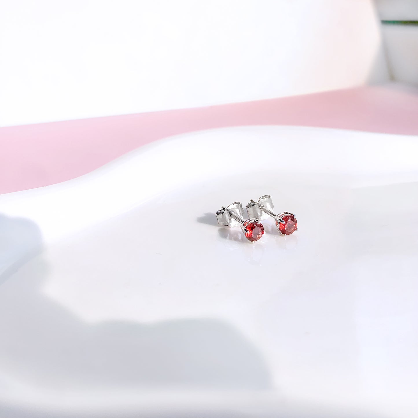 Close-up front angle of a pair of white metal stud earrings with round red gemstones in four-prong settings placed on a white curved surface.