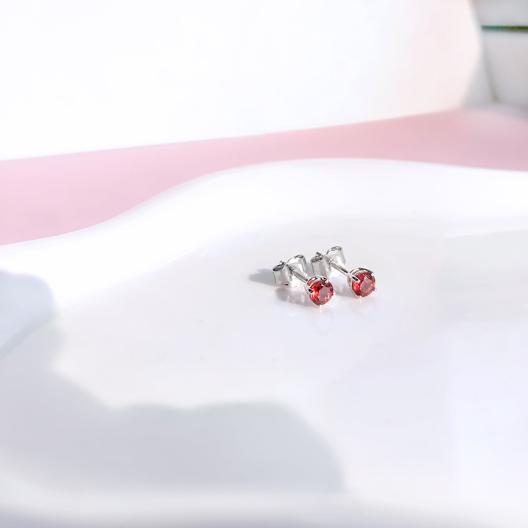 Close-up front angle of a pair of white metal stud earrings with round red gemstones in four-prong settings placed on a white curved surface.