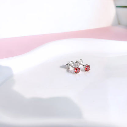 Close-up front angle of a pair of white metal stud earrings with round red gemstones in four-prong settings placed on a white curved surface.