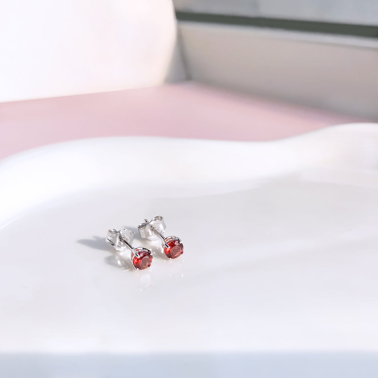 Pair of round red gemstone stud earrings in four-prong white settings shown from a slightly angled top view on a white surface.