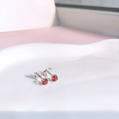Pair of round red gemstone stud earrings in four-prong white settings shown from a slightly angled top view on a white surface.