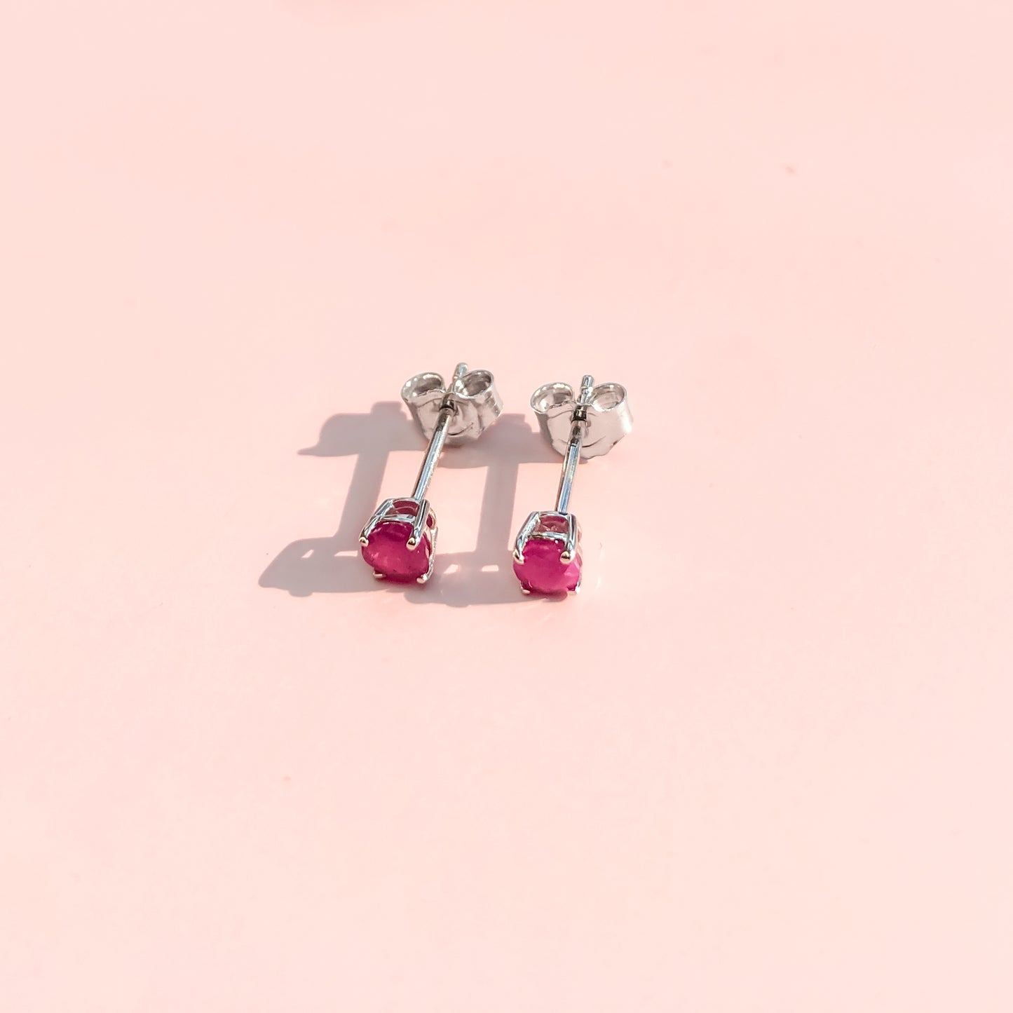 Pair of white metal stud earrings with round pink gemstones in 4-prong settings, shown from a top front angle on a pink background.