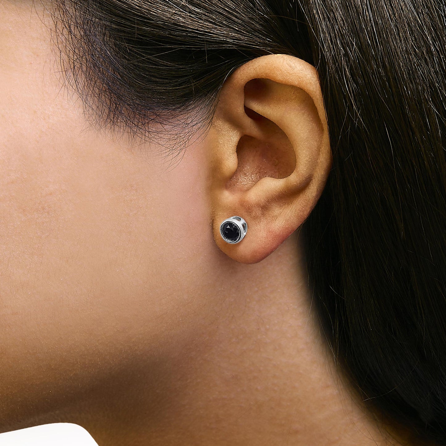 Close-up side view of a model wearing a pair of round white metal stud earrings, each set with a black round gemstone in a bezel setting.