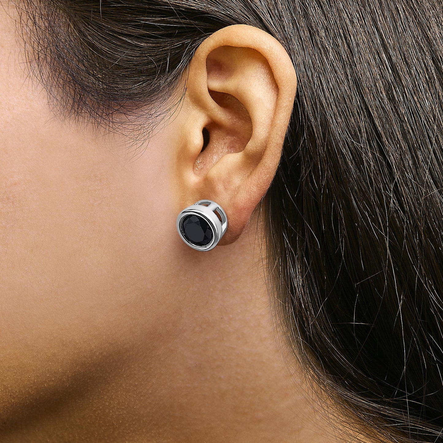 Close-up side view of a white metal round bezel-set black gemstone stud earring shown worn on a model's ear.