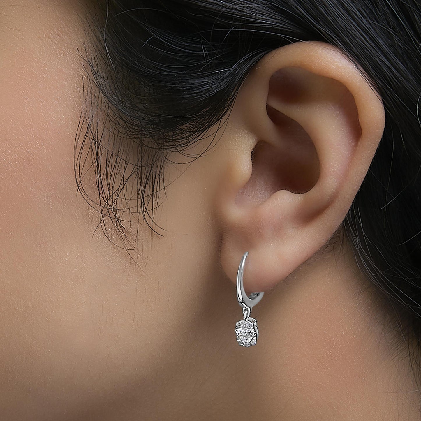 Close-up front angle of a white metal earring worn on a model’s ear, featuring a small hoop with a dangling round cluster of white gemstones in prong settings.