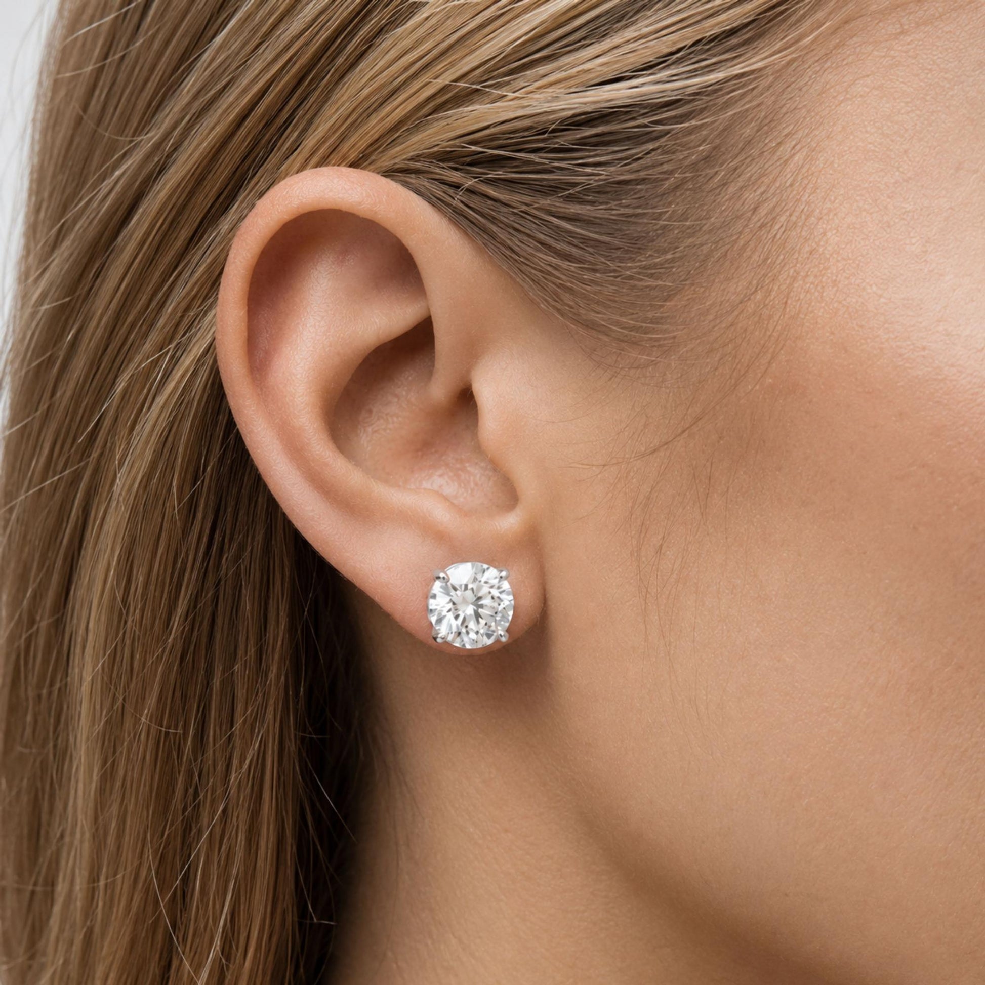 Close-up side view of a model wearing a white metal stud earring with a round white gemstone in a four-prong setting.
