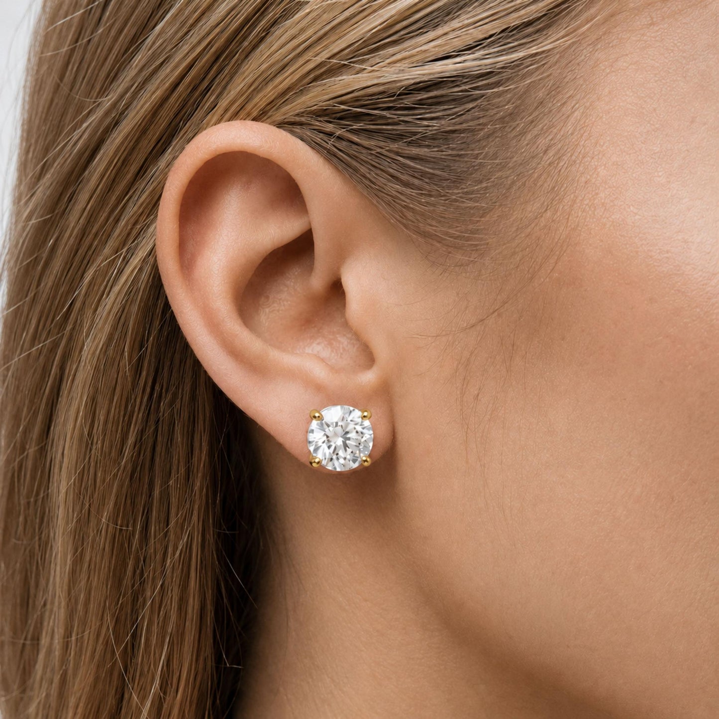 Close-up side view of a model wearing a yellow metal stud earring with a round white gemstone in a four-prong setting.