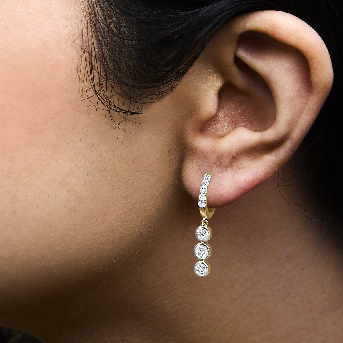 Close-up side view of a model wearing yellow metal earrings featuring a curved design with round white gemstones in prong and bezel settings, and three dangling bezel-set round white gemstones.