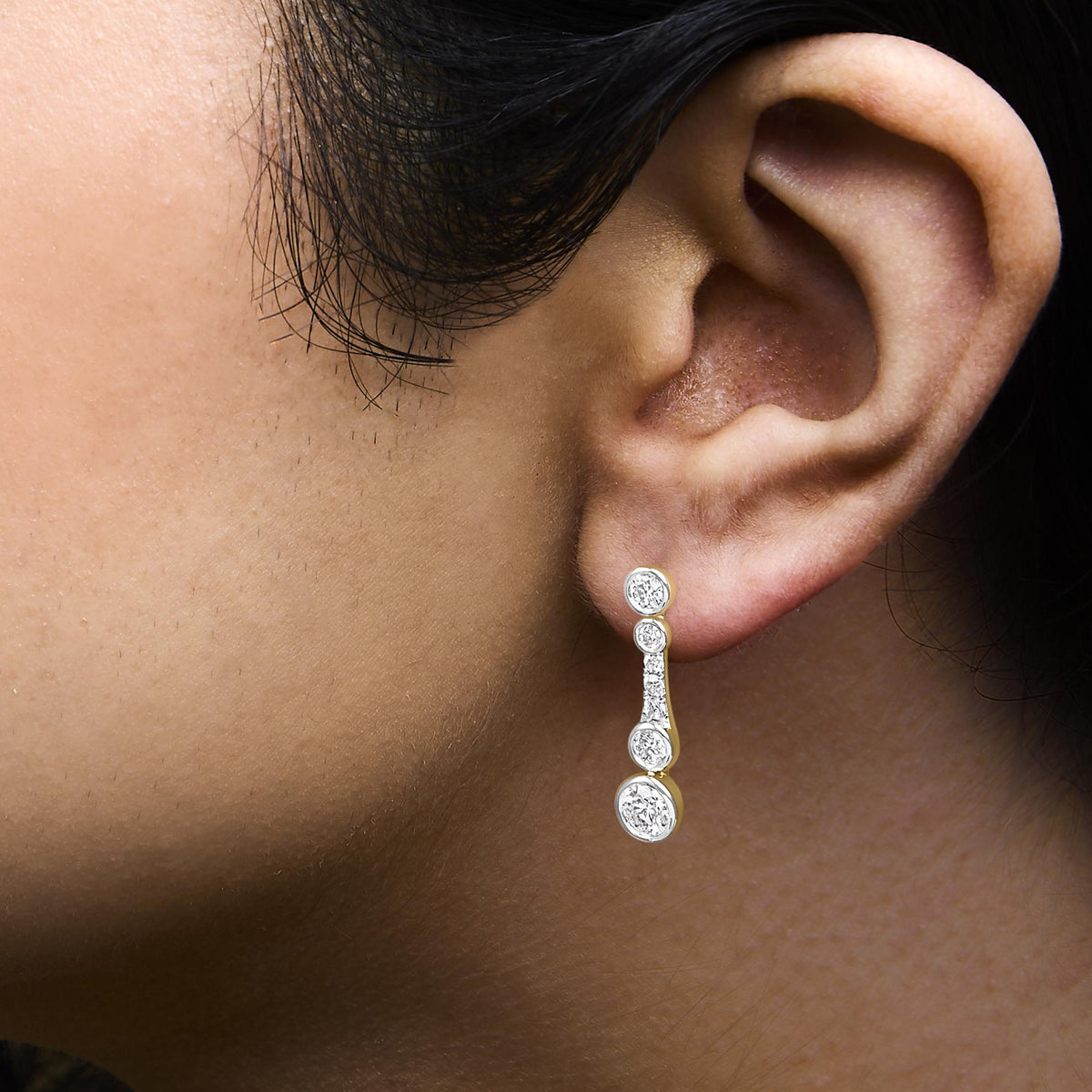 Close-up side view of a model’s ear wearing a yellow metal earring featuring multiple round white gemstones in bezel and prong settings arranged vertically.