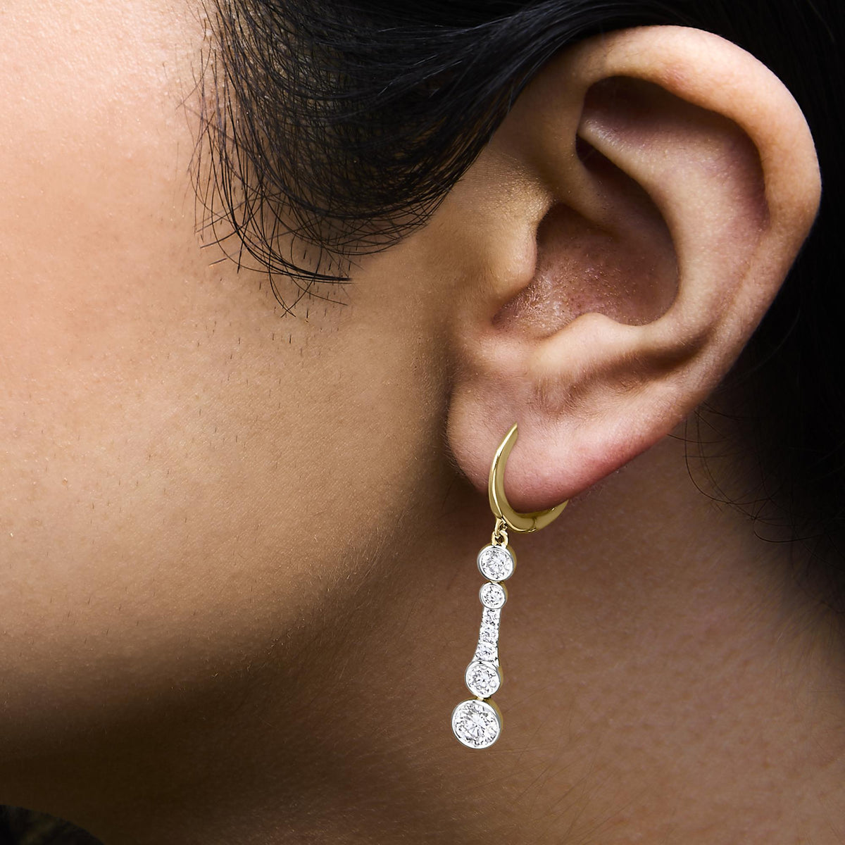 Close-up side view of a model wearing a yellow metal earring featuring a curved hoop with multiple round white gemstones in bezel and prong settings dangling from it.