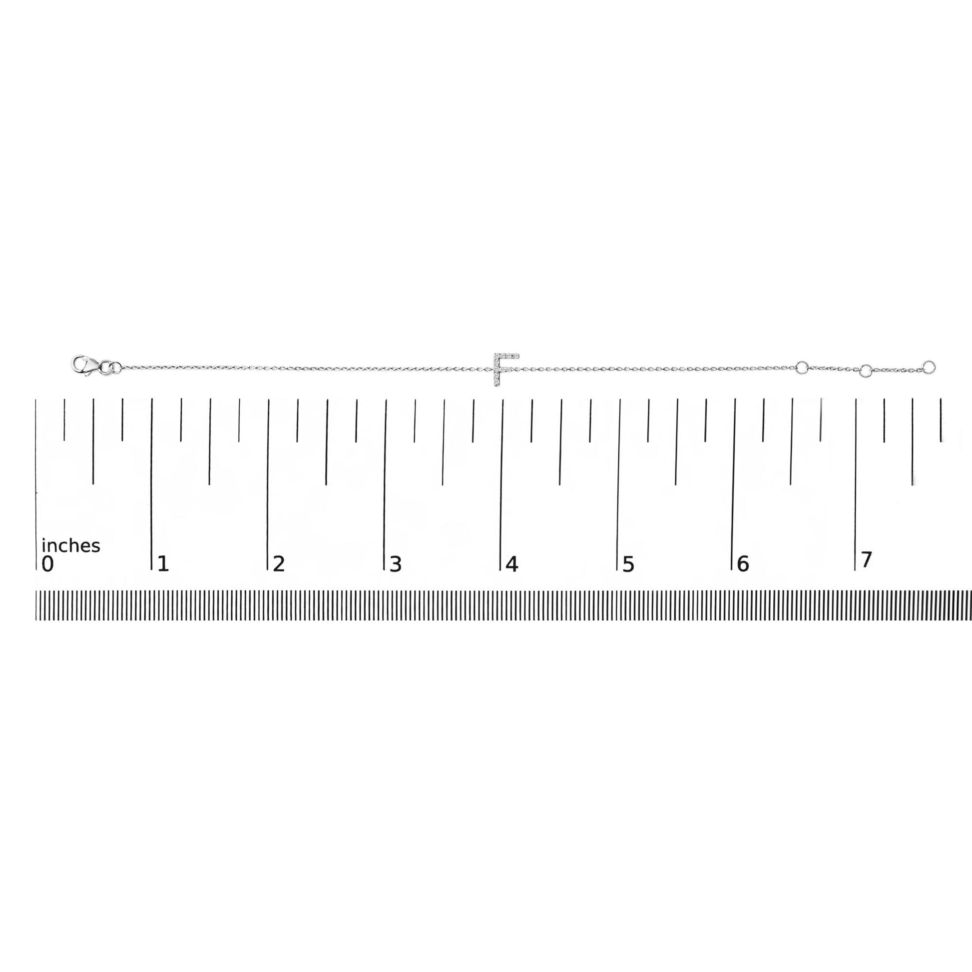White metal bracelet with a letter 'F' featuring white gemstones in a micro-pave setting, shown lying flat on a measuring scale from a top-down angle.