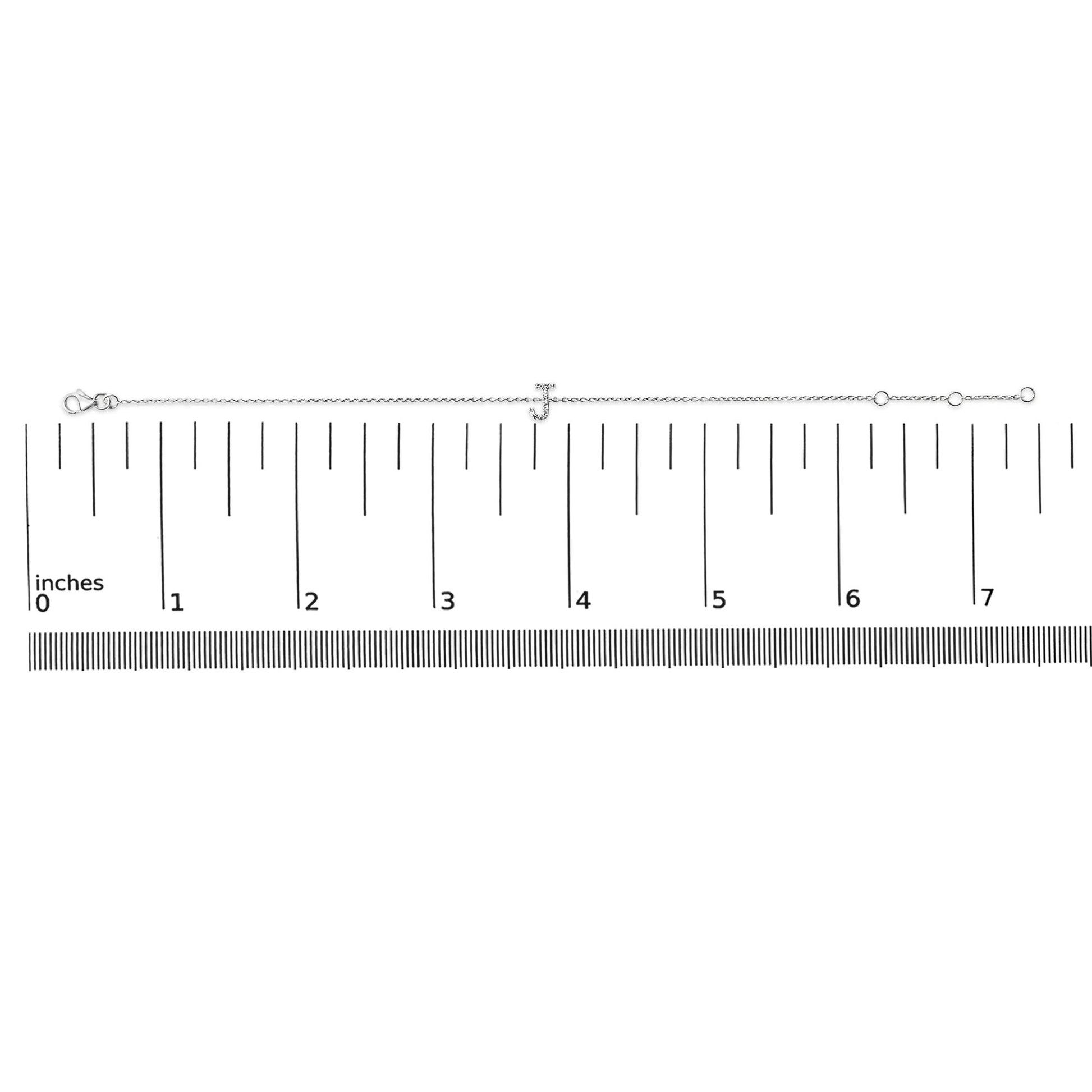 Bracelet with a white metal chain and a letter 'J' centerpiece adorned with small white gemstones, shown flat on a ruler scale from a top-down angle.