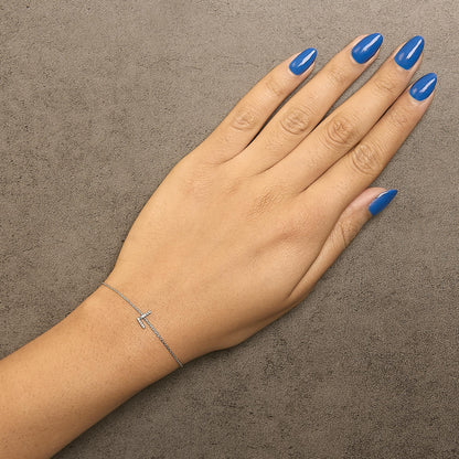 White delicate bracelet with a small T-shaped detail featuring round white gemstones in micro-pave setting, shown worn on a model's wrist with blue nail polish, top-down angle.