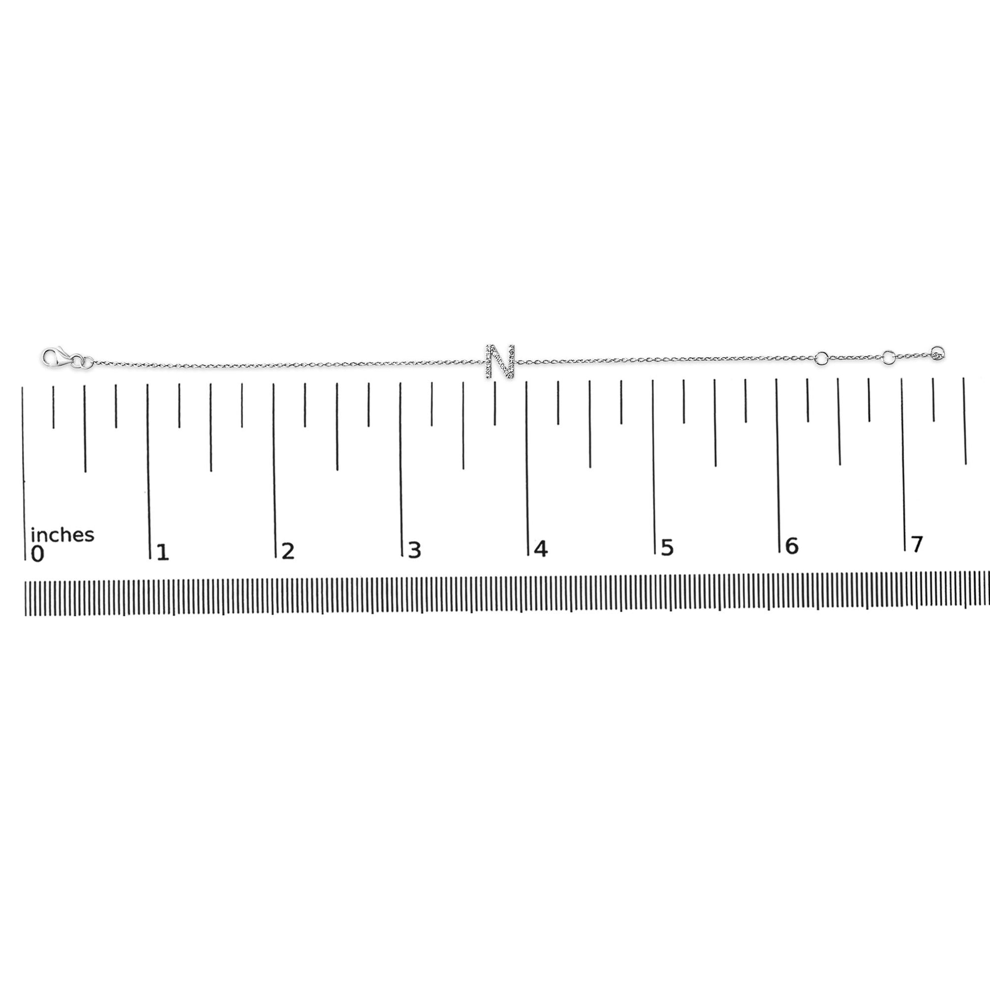 White metal bracelet with an 'N' letter centerpiece adorned with small white gemstones in a micro-pave setting, laid flat on a horizontal ruler, top-down angle.