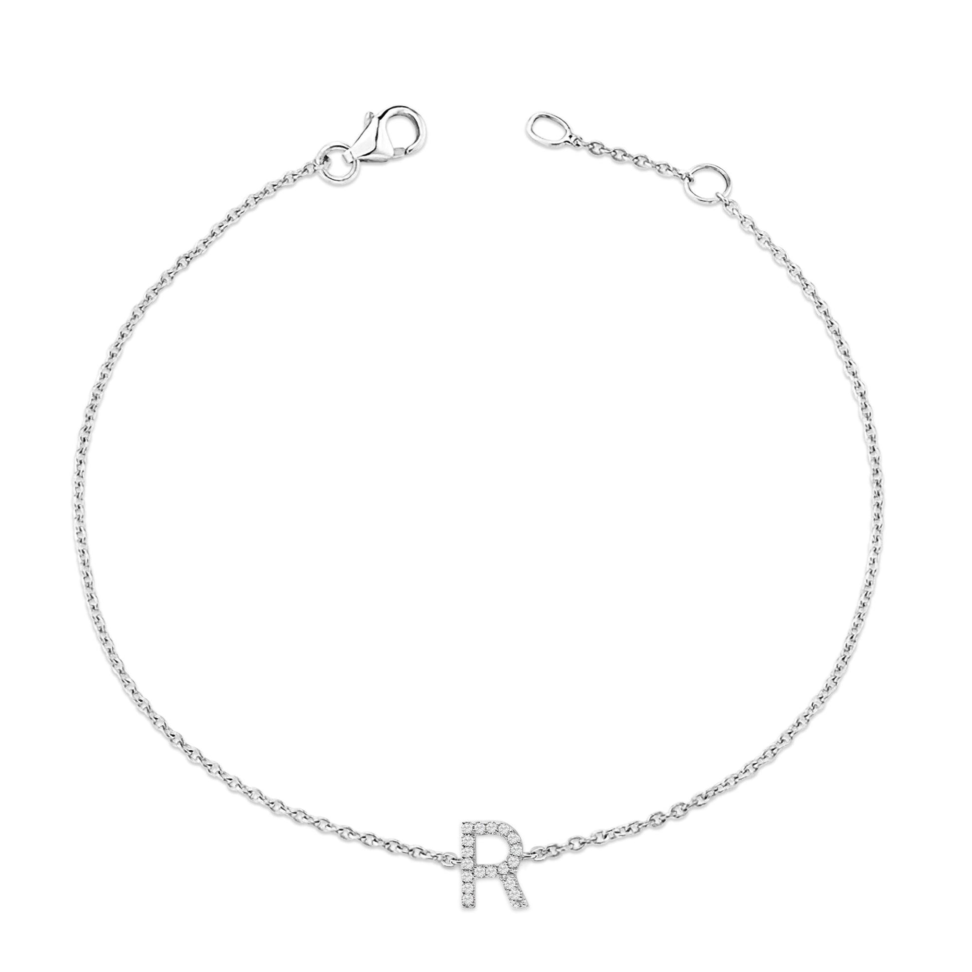 Top-down view of a delicate white bracelet chain with a letter 'R' charm adorned with small white round gemstones in a micro-pave setting, shown unclasped on a white background.