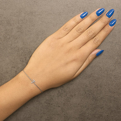 Bracelet with a small T-shaped charm set with white gemstones on a delicate white chain, shown on a model's wrist from a top-down angle.