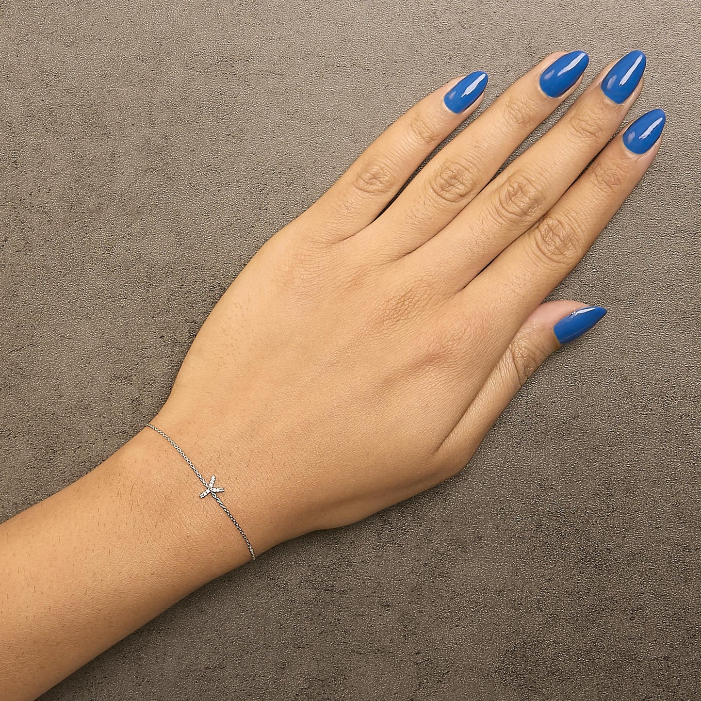 White metal bracelet with a small cross-shaped detail featuring round white gemstones in a micro-pave setting, shown worn on a model's wrist from a top-down angle.