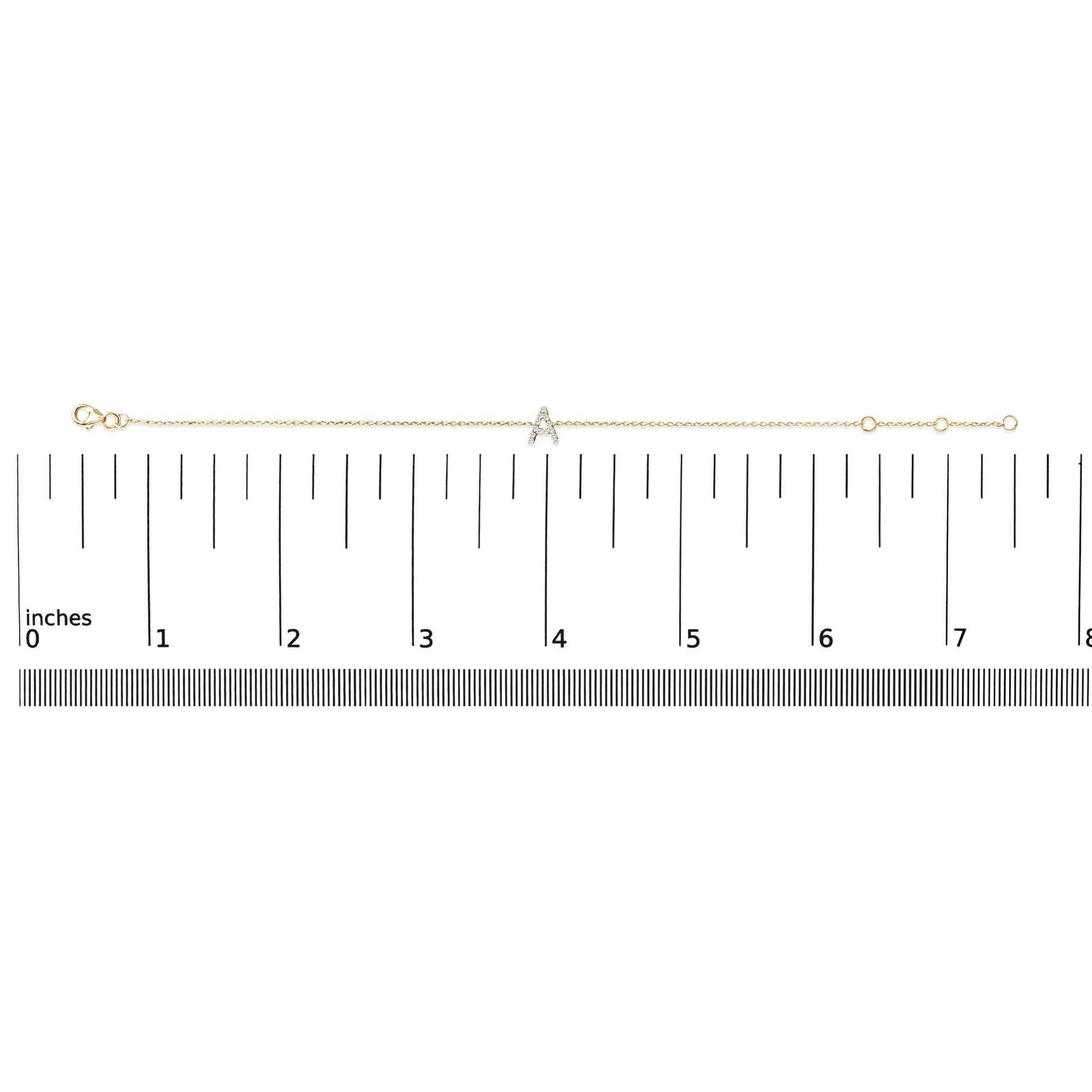 Yellow metal bracelet with a small 'A' shaped charm set with white gemstones in micro-pave setting, displayed horizontally on a ruler scale, top-down view.