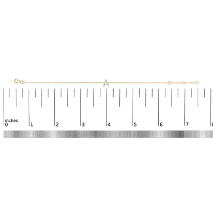 Yellow metal bracelet with a small 'A' shaped charm set with white gemstones in micro-pave setting, displayed horizontally on a ruler scale, top-down view.