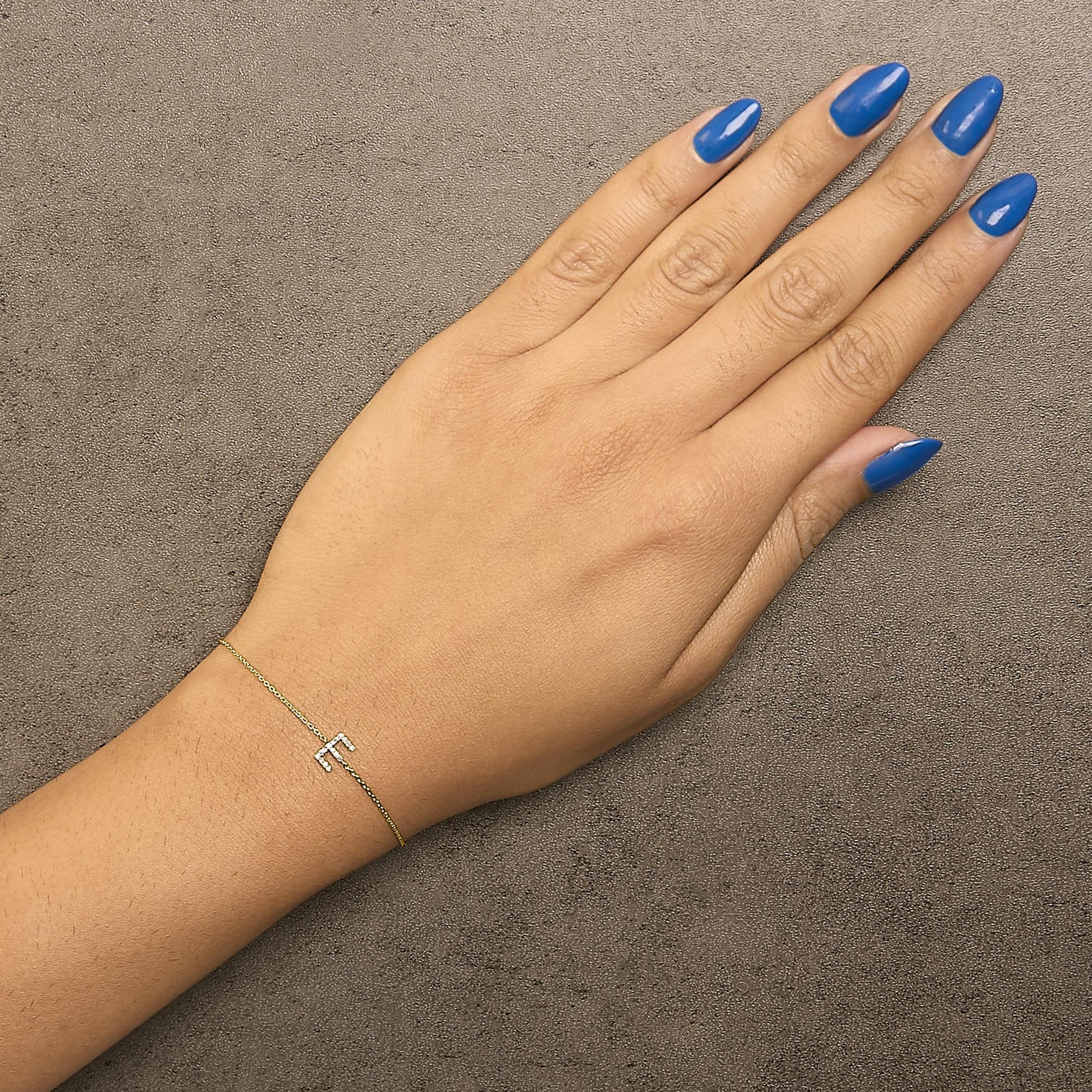 Bracelet with a yellow chain and a small letter 'F' adorned with white round gemstones in micro-pave setting, shown worn on a model's wrist from a top-down angle.