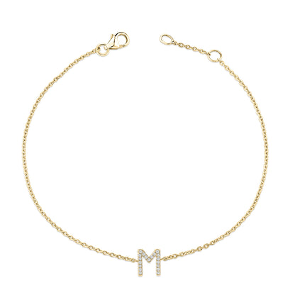 Top-down view of a yellow metal bracelet with a chain and an 'M' shaped charm featuring round white gemstones in a micro-pave setting.